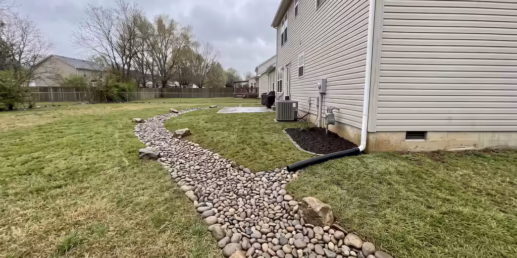After: The same yard with the downspout piped underground, a river-rock channel through the low spot, regraded lawn and fresh sod