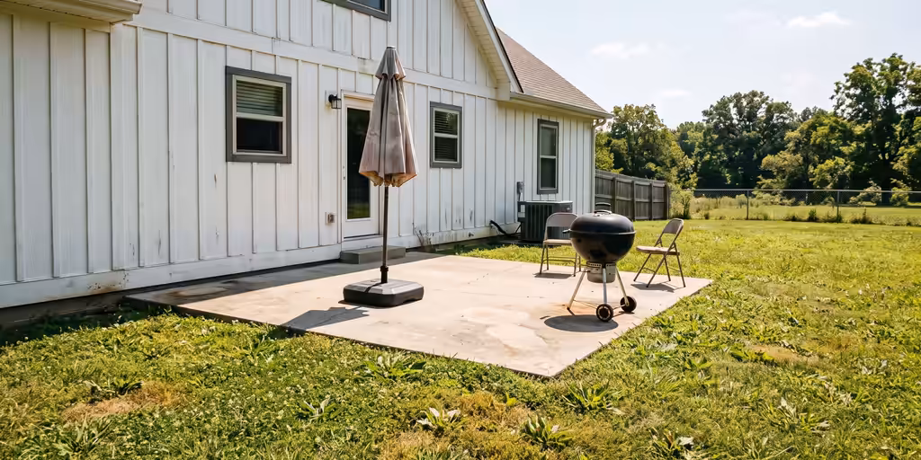 Before: Plain concrete patio in full sun behind a white farmhouse-style home, with an umbrella and a grill