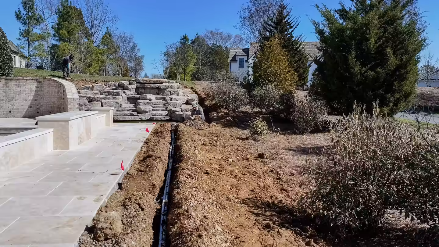 Drain line trench running beside a new travertine terrace and boulder retaining wall