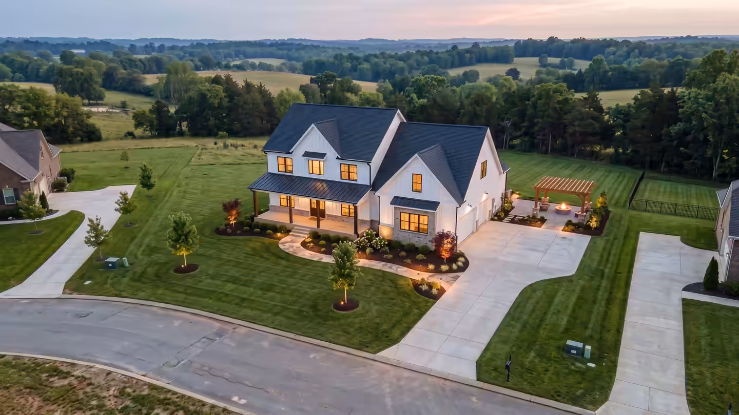 Concept image: drone view of a newly landscaped farmhouse at dusk, with lit walk, planted beds, fresh sod and a fire under the patio pergola