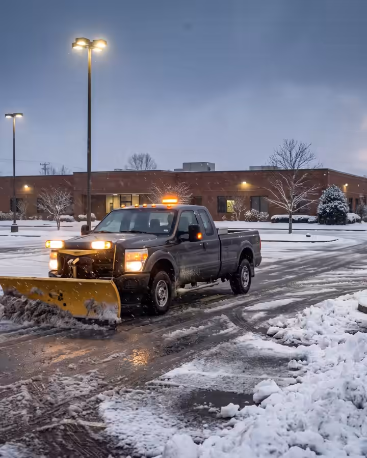 Pickup with a plow clearing a commercial parking lot at dawn